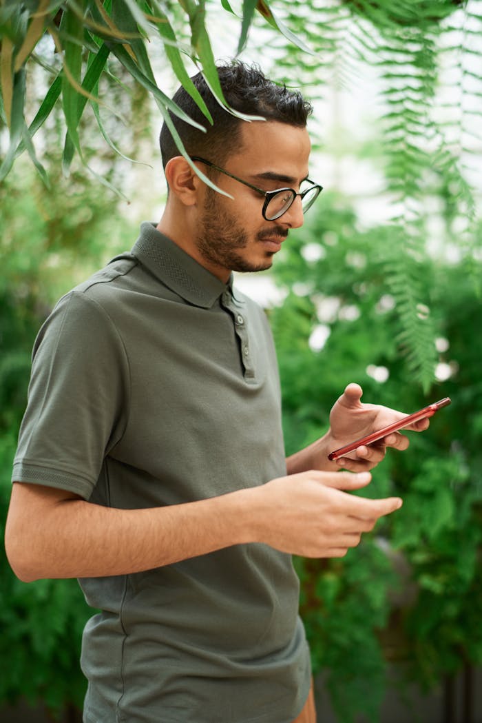Young man checks phone surrounded by lush green plants, evoking a natural setting.